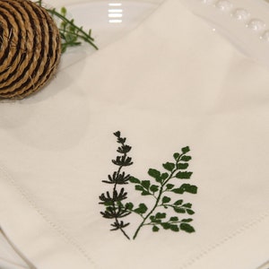 Puede incluir: Servilleta de lino blanco con un dise&ntilde;o de helecho bordado en verde y negro.