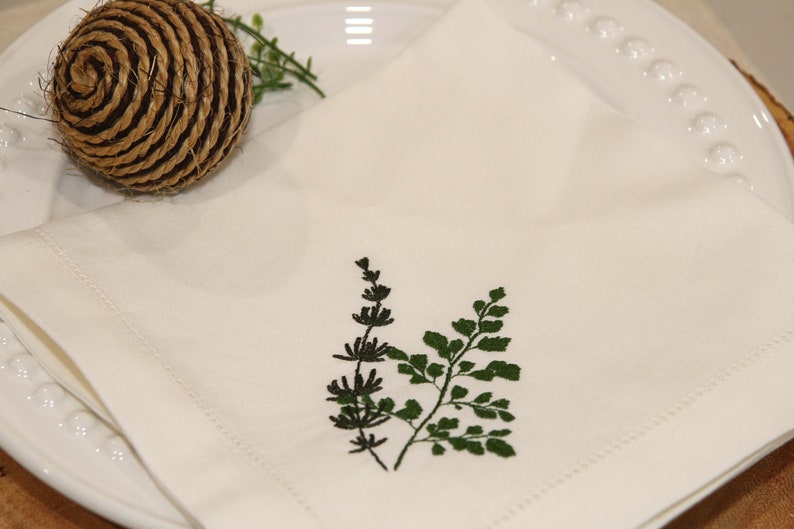 Puede incluir: Servilleta de lino blanco con un dise&ntilde;o de helecho bordado en verde y negro.