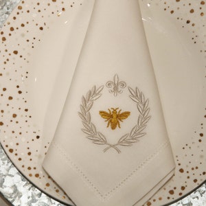 May include: A white linen napkin with a gold bee and silver laurel wreath embroidery. The napkin is folded and placed on a white plate with gold speckles.