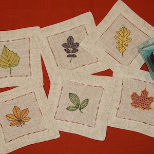 Peut inclure: Ensemble de six sous-verres en lin avec des feuilles d'automne brodées dans différentes couleurs et motifs. Les sous-verres sont beiges avec une bordure rouge et présentent une feuille de bouleau jaune, une feuille de chêne violette, une feuille d'érable verte, une feuille de chêne jaune, une feuille d'érable rouge et une feuille verte.
