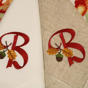 May include: Two embroidered napkins with the letter "B" in red, surrounded by fall leaves and acorns. The napkins are made of different fabrics, one white and one beige.