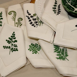 Puede incluir: Un juego de servilletas de lino blanco con dise&ntilde;os de helechos verdes bordados. Las servilletas est&aacute;n dobladas y dispuestas sobre una superficie beige.