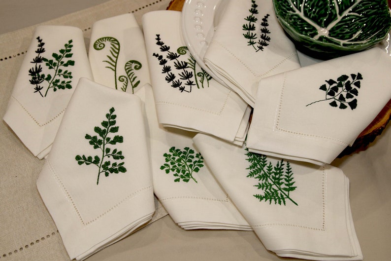 Puede incluir: Un juego de servilletas de lino blanco con dise&ntilde;os de helechos verdes bordados. Las servilletas est&aacute;n dobladas y dispuestas sobre una superficie beige.