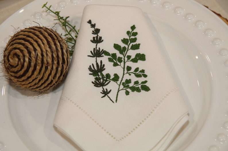 Puede incluir: Servilleta de lino blanco con un dise&ntilde;o de helecho y hojas bordadas en verde.