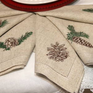 May include: Set of five beige linen napkins with embroidered pine cone designs in shades of brown and green.