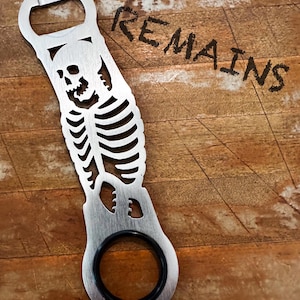 May include: A silver bottle opener with a skeleton design. The opener features a skull and rib cage cut-out design. The word "REMAINS" is written in black on the wooden background. The opener has a black rubber ring.