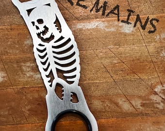 May include: A silver bottle opener with a skeleton design. The opener features a skull and rib cage cut-out design. The word "REMAINS" is written in black on the wooden background. The opener has a black rubber ring.