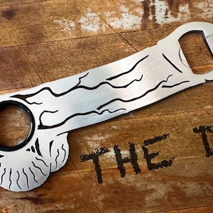 May include: A silver metal bottle opener with a unique design. The opener features a circular opening with a black interior and a bottle cap opener at the end. The words "THE D" are stenciled on the wooden surface.