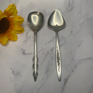 May include: Two silver spoons with ornate handles. The spoon on the left has a rounded bowl, while the spoon on the right has a pointed bowl.
