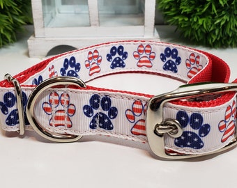 May include: A red and white dog collar with a patriotic design. The collar features a white ribbon with blue and red paw prints, and a red backing. The collar has a silver metal buckle and a D-ring.