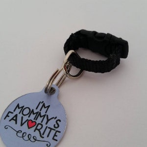 May include: A light blue pet tag with the text "I'M MOMMY'S FAVORITE" in black and a red heart. The tag is attached to a black nylon strap with a black plastic buckle and a silver metal ring.