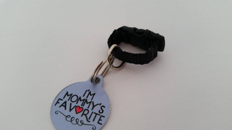 May include: A light blue pet tag with the text "I'M MOMMY'S FAVORITE" in black and a red heart. The tag is attached to a black nylon strap with a black plastic buckle and a silver metal ring.