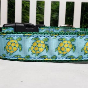 May include: A dog collar with a light blue background featuring a pattern of yellow and green turtles. The collar has a green trim and a black plastic buckle. The collar is approximately 1 inch wide.