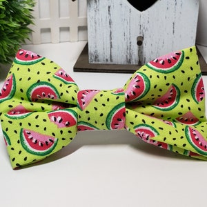May include: A bow tie in a vibrant green fabric, patterned with watermelon slices. The watermelons feature red and pink segments, green rinds, and black seeds. The bow tie is a traditional shape, likely made of cloth.