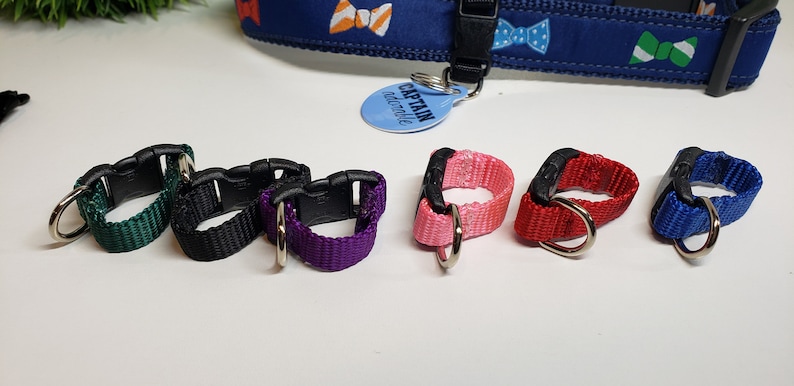 May include: A collection of colorful dog collar accessories, including green, black, purple, pink, red, and blue loops with metal D-rings. A blue collar with bow tie patterns is visible in the background.