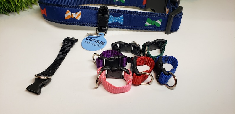 May include: A blue dog collar with bow tie patterns, a black leash attachment, and several colored collar attachments. A light blue tag reads "CAPTAIN adorable".