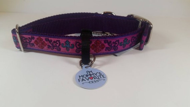 May include: A purple dog collar with a pink and patterned design. The collar has a metal buckle and a small tag that reads "I'M MOMMY'S FAVORITE". The collar is designed for a pet.