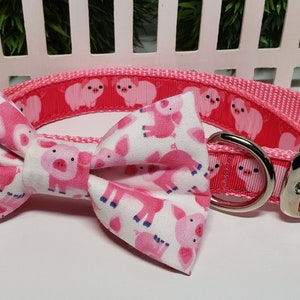 May include: Pink dog collar and bow tie set. The collar is pink with a repeating pattern of white pig illustrations. The bow tie is white with a pink pig pattern. The collar has a silver metal buckle.