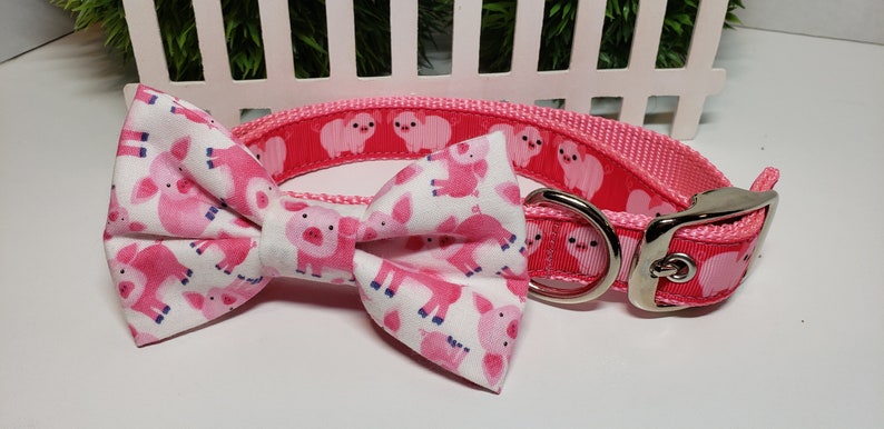 May include: Pink dog collar and bow tie set. The collar is pink with a repeating pattern of white pig illustrations. The bow tie is white with a pink pig pattern. The collar has a silver metal buckle.