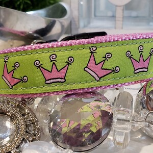 May include: A dog collar with a lime green band featuring pink crown graphics. The collar has a pink trim and a black buckle. The image is set against a backdrop of decorative objects.