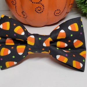 May include: A black bow tie with a repeating pattern of candy corn in orange, yellow, and white. The bow tie is centered in the image, with a pumpkin and greenery in the background. The candy corn pattern is a festive Halloween design.