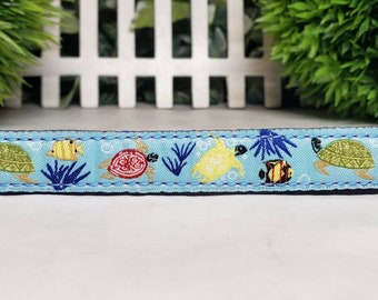 May include: Light blue pet collar featuring a repeating pattern of sea turtles, fish, and coral. The design includes yellow, green, red, and blue elements. The collar has a black underside and is bordered with blue stitching.