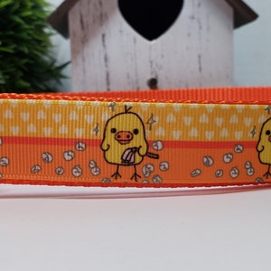 May include: Orange dog collar featuring a repeating pattern of yellow chicks with sticks, set against a yellow and orange background. The collar has a bright orange plastic buckle. The design includes white heart shapes and small white circles.