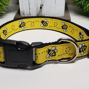 May include: A yellow dog collar with a black trim and a black plastic buckle. The collar features a repeating pattern of cartoon bees with white wings and black outlines. The collar has a silver metal D-ring.