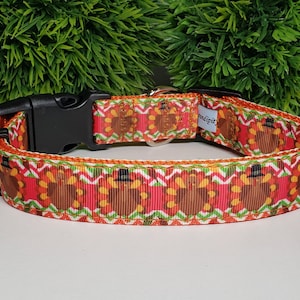 May include: A festive dog collar featuring a repeating pattern of cartoon turkeys in top hats on a red background. The collar has a black plastic buckle and a white and green zig-zag border. The collar is designed for pets.