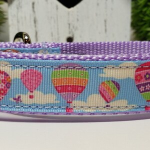 May include: A dog collar with a light blue background featuring colorful hot air balloons, white clouds, and a purple trim. The collar has a metal clasp and a matching purple nylon strap.