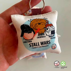 Può includere: Un portachiavi in tessuto bianco con personaggi dei cartoni animati di "Stall Wars". Il design include "Lard Fener", "Chewvacca" e "Can Solo". Il portachiavi ha un anello di metallo e un passante in tessuto bordeaux.