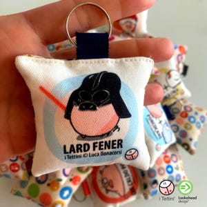 Può includere: Un portachiavi quadrato in tessuto raffigurante un maialino dei cartoni animati che indossa un casco nero e tiene una spada laser rossa. Il testo LARD FENER è stampato sotto il personaggio, con i Tettini Luca Bonacorsi sotto. Anello incluso.