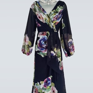 Puede incluir: Un vestido formal azul marino de manga larga con un estampado floral en tonos rosas, morados y verdes. El vestido presenta un escote en V y un corpi&ntilde;o cruzado. Un tocado verde a juego y un collar de perlas completan el conjunto.