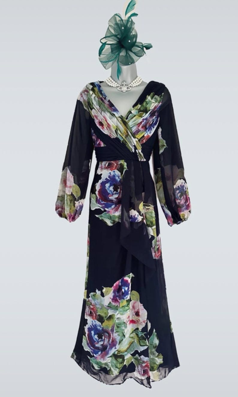 Puede incluir: Un vestido formal azul marino de manga larga con un estampado floral en tonos rosas, morados y verdes. El vestido presenta un escote en V y un corpi&ntilde;o cruzado. Un tocado verde a juego y un collar de perlas completan el conjunto.