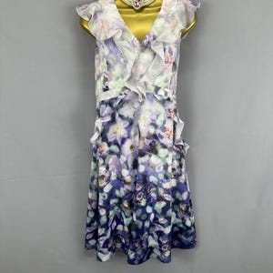 May include: A sleeveless floral print dress with a V-neckline and ruffled trim. The dress features a watercolor-style floral pattern in shades of purple, blue, and white. A white beaded choker necklace with a pink heart-shaped pendant is also visible.