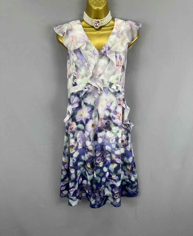 May include: A sleeveless floral print dress with a V-neckline and ruffled trim. The dress features a watercolor-style floral pattern in shades of purple, blue, and white. A white beaded choker necklace with a pink heart-shaped pendant is also visible.
