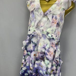May include: A sleeveless floral print dress in shades of purple, white, and green. The dress features a V-neckline, ruffled accents, and a flowing skirt. A pearl and pink gem choker necklace is also visible.