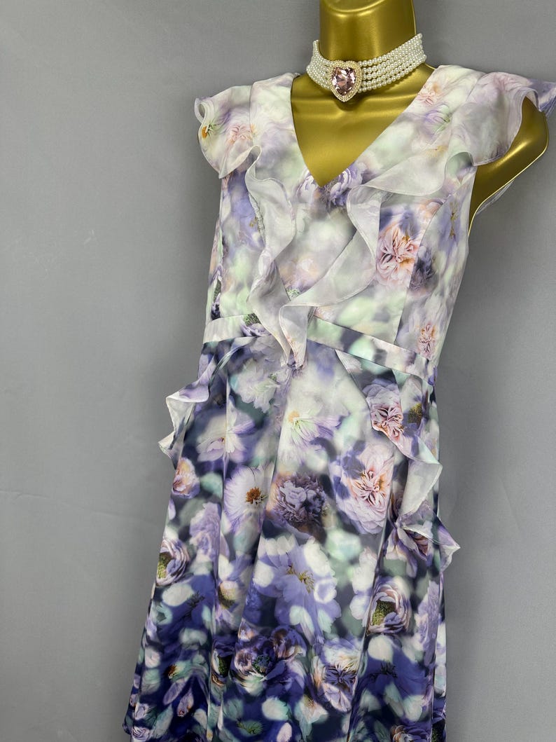 May include: A sleeveless floral print dress in shades of purple, white, and green. The dress features a V-neckline, ruffled accents, and a flowing skirt. A pearl and pink gem choker necklace is also visible.