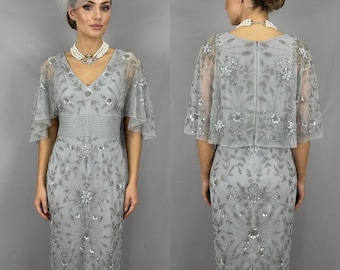 May include: A knee-length silver-grey dress featuring a V-neckline, short flutter sleeves, and a fitted waist. The dress is adorned with intricate silver sequin and bead embroidery in a floral pattern, complemented by a matching fascinator headpiece.