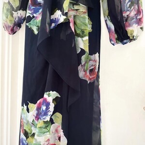 Puede incluir: Vestido largo negro con estampado floral. El vestido presenta un detalle de tela en cascada en la parte delantera y mangas largas. El estampado floral incluye flores rosas, moradas, azules y verdes.