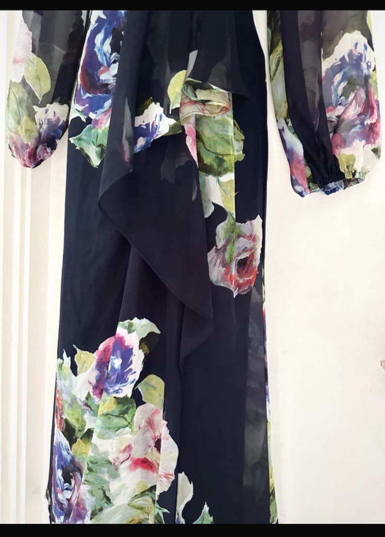 Puede incluir: Vestido largo negro con estampado floral. El vestido presenta un detalle de tela en cascada en la parte delantera y mangas largas. El estampado floral incluye flores rosas, moradas, azules y verdes.