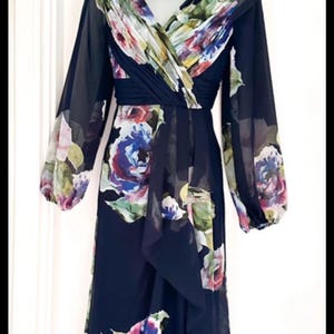 Puede incluir: Vestido azul marino de manga larga con estampado floral en tonos morados, azules y blancos. El vestido presenta un escote en V y una falda fluida. Un fascinador verde oscuro a juego y un collar de perlas completan el conjunto.