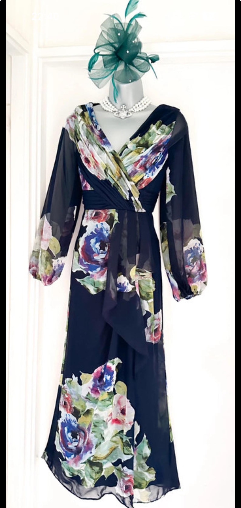 Puede incluir: Vestido azul marino de manga larga con estampado floral en tonos morados, azules y blancos. El vestido presenta un escote en V y una falda fluida. Un fascinador verde oscuro a juego y un collar de perlas completan el conjunto.