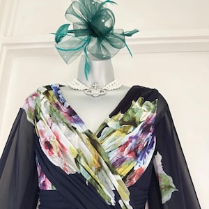 Puede incluir: Un vestido azul marino oscuro con un corpi&ntilde;o estampado floral y mangas transparentes. El vestido est&aacute; adornado con un fascinador verde y un collar de perlas con un colgante plateado. El vestido se exhibe en un maniqu&iacute;.
