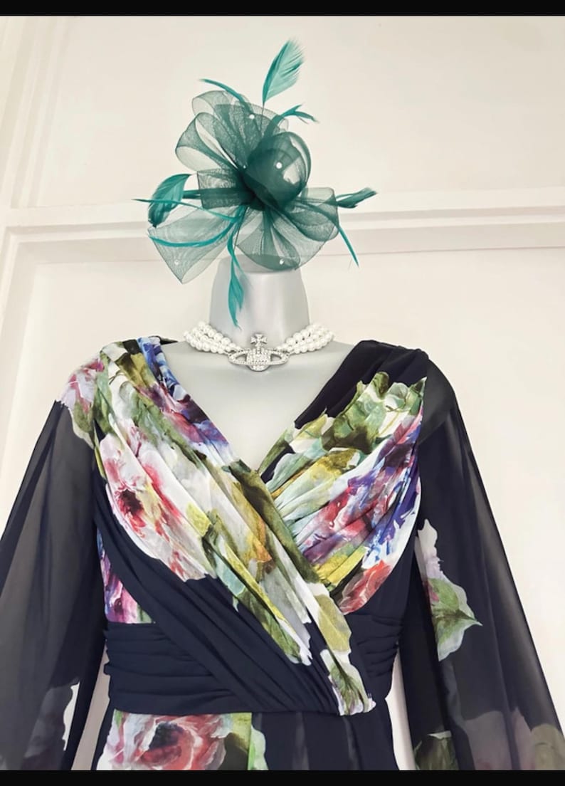 Puede incluir: Un vestido azul marino oscuro con un corpi&ntilde;o estampado floral y mangas transparentes. El vestido est&aacute; adornado con un fascinador verde y un collar de perlas con un colgante plateado. El vestido se exhibe en un maniqu&iacute;.