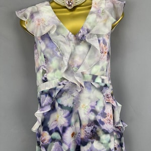 May include: A floral print dress with a V-neck and ruffled trim. The dress features a pattern of purple, white, and green flowers. A pearl choker with a pink heart-shaped pendant is worn with the dress.