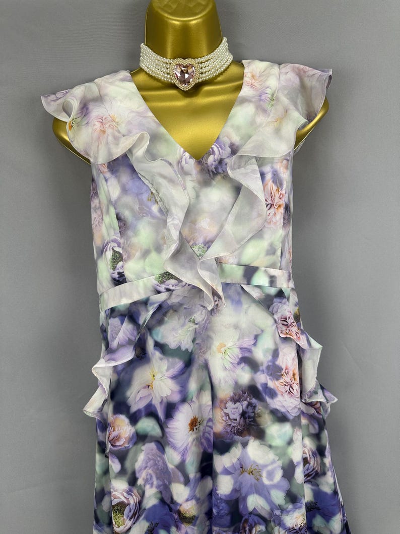 May include: A floral print dress with a V-neck and ruffled trim. The dress features a pattern of purple, white, and green flowers. A pearl choker with a pink heart-shaped pendant is worn with the dress.