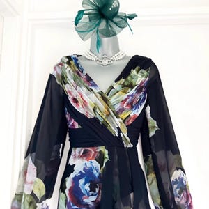 Puede incluir: Vestido formal con estampado floral, con mangas largas transparentes y escote en V. El vestido se combina con un tocado verde y un collar de perlas. El estampado floral incluye tonos de azul, morado y rosa.