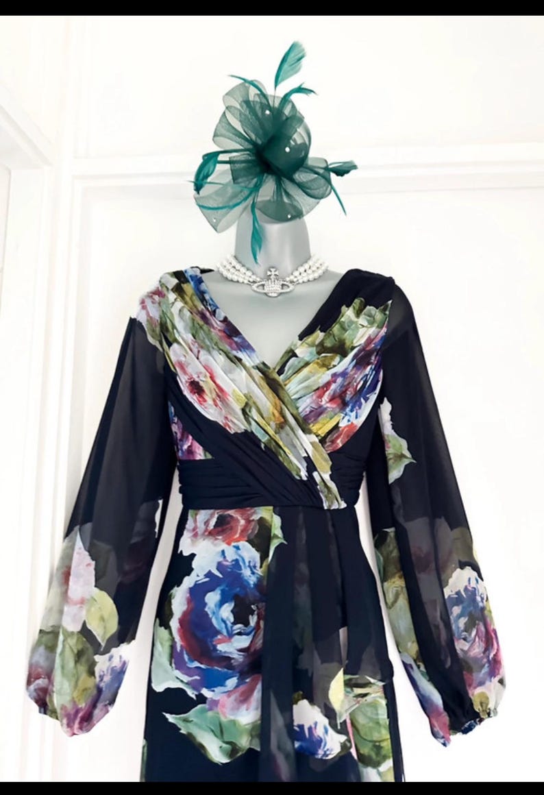 Puede incluir: Vestido formal con estampado floral, con mangas largas transparentes y escote en V. El vestido se combina con un tocado verde y un collar de perlas. El estampado floral incluye tonos de azul, morado y rosa.