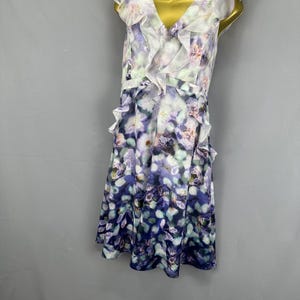 May include: A sleeveless floral print dress with a V-neck and ruffled trim. The dress features a white, purple, and blue floral pattern. A white choker necklace with a pink heart-shaped pendant is also visible.